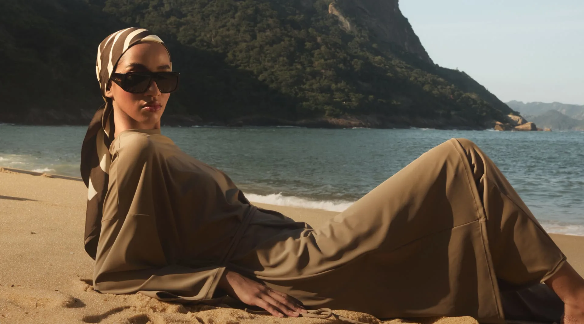 A Merrachi campaign frame: a model in olive-toned modest tailoring and a printed silk headscarf reclining on the sand, a forested headland behind her.